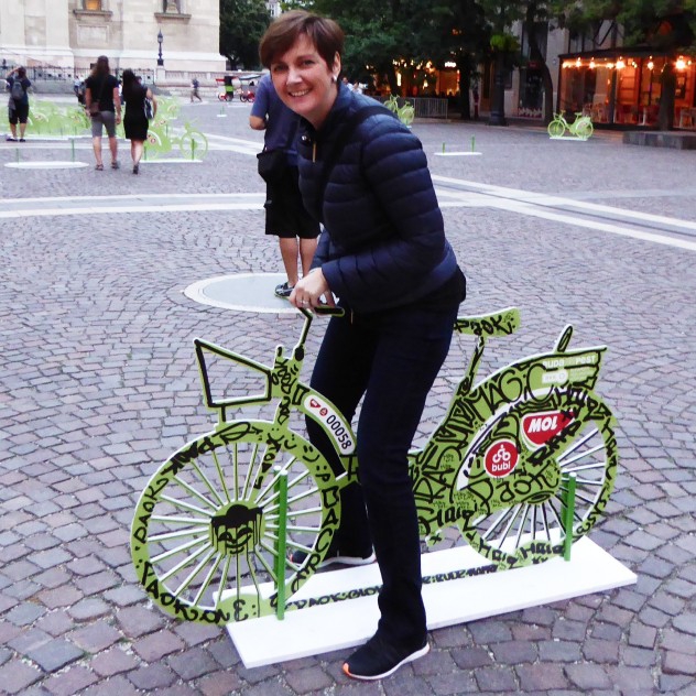Natalie posing on a bicycle sculpture in a cobblestoned square.