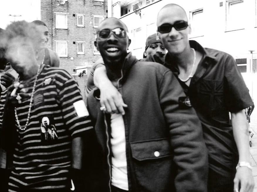A black and white image of So Solid Crew, with one member wearing a Stone Island jacket. / Image en noir et blanc de So Solid Crew, dont l’un des membres est vêtu d’une veste Stone Island.
