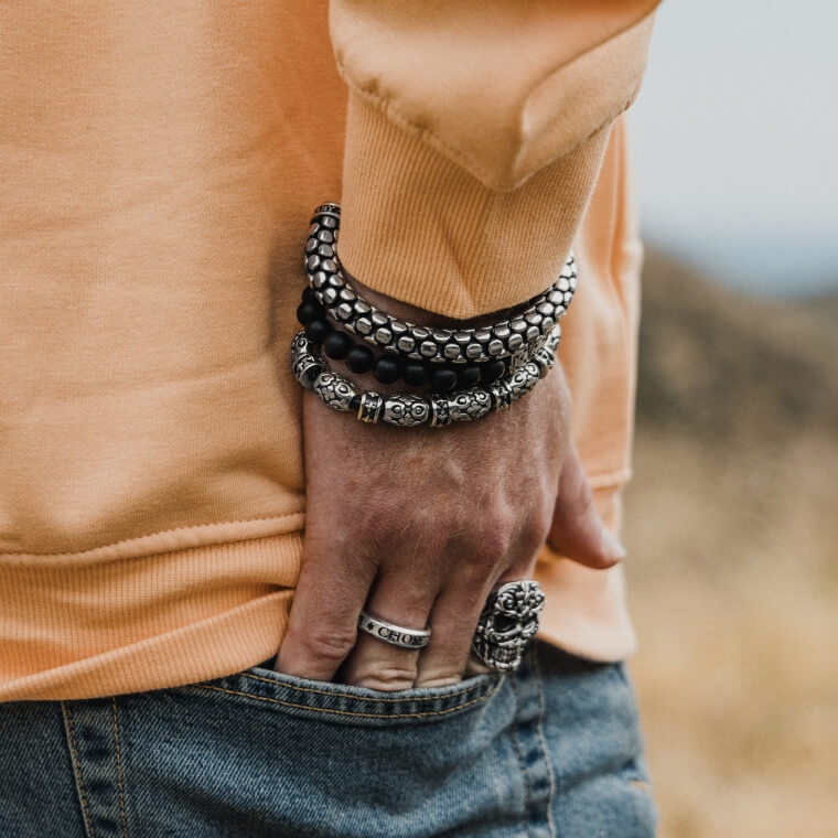 A close up of a male model wearing rings and bracelets, tucking his hand into his back jean’s pocket.