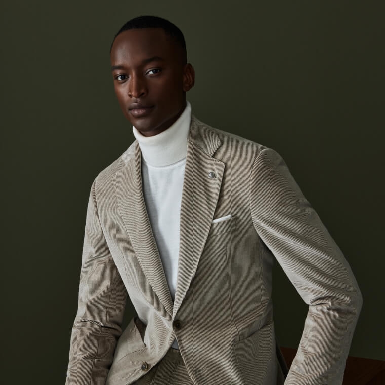 A male model wearing a grey corduroy suit and white turtleneck.